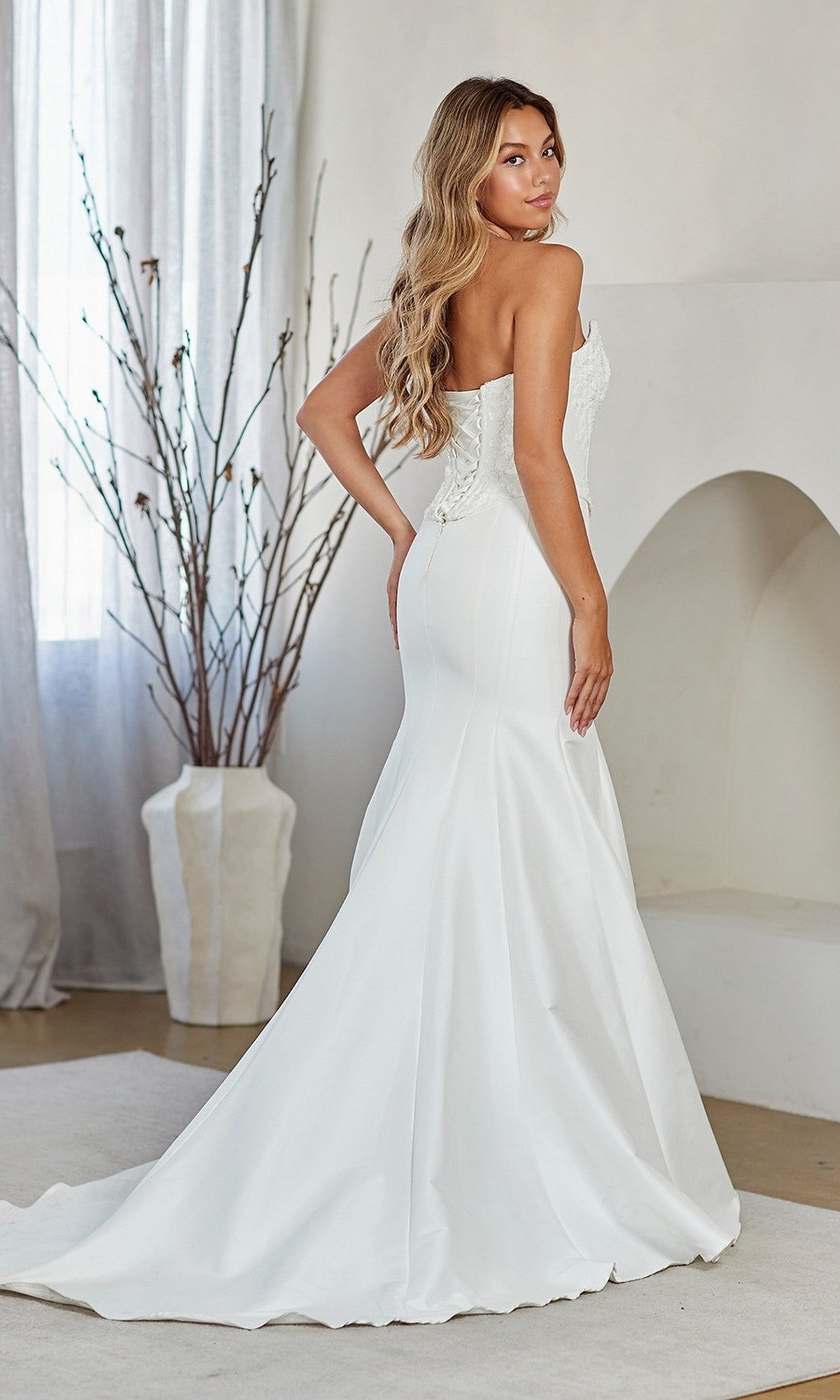 A woman wears the Amelia Couture 8080 Long Prom Dress—a strapless white gown with an embellished corset bodice and fit-and-flare skirt—standing indoors, facing slightly away from the camera.